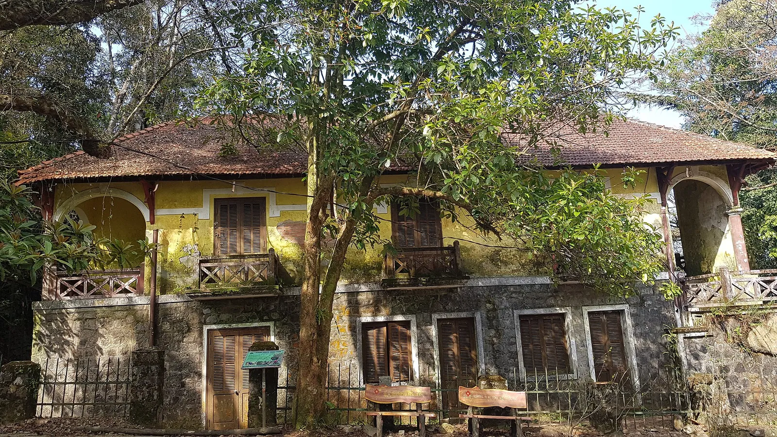 A two-storey French colonial villa with yellow walls, a tiled roof and arched verandas among trees under a blue sky in Bach Ma National Park