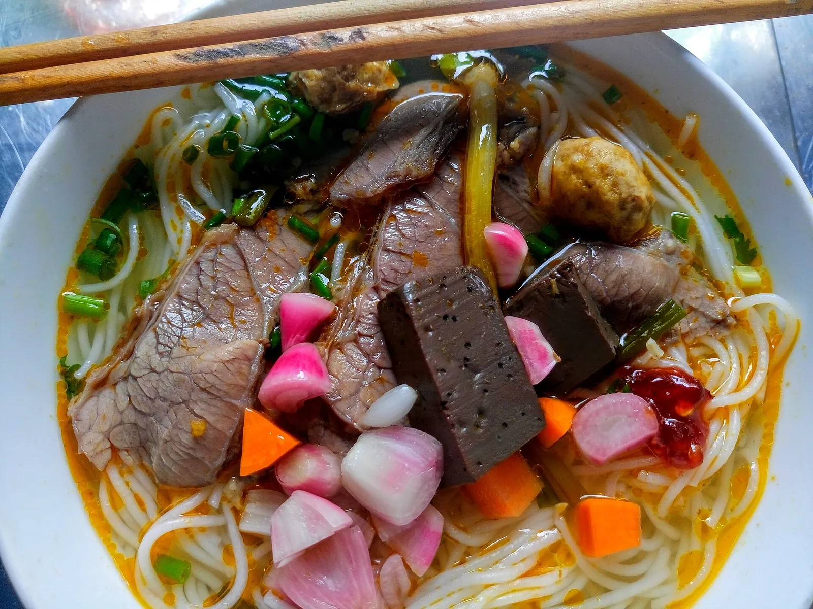 A bowl of bun bo Hue with round rice noodles, slices of beef, blood cubes, pork, herbs and chilli in a red broth, chopsticks resting on the rim