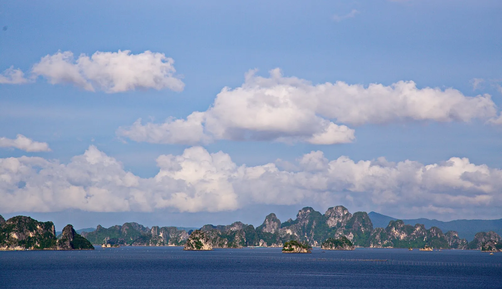 A long line of forested limestone islands on blue water under a bright sky in Bai Tu Long Bay