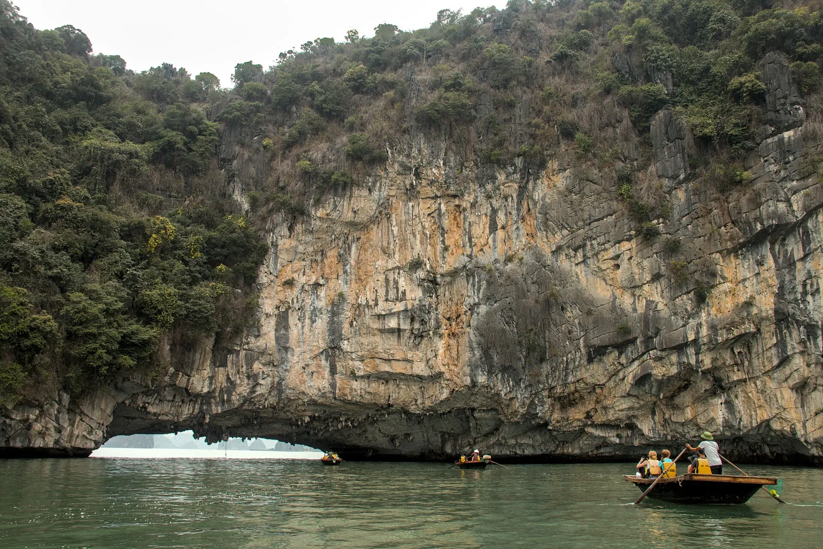 A small rowing boat with visitors passing a limestone cliff with a natural arch at sea level in Bai Tu Long Bay