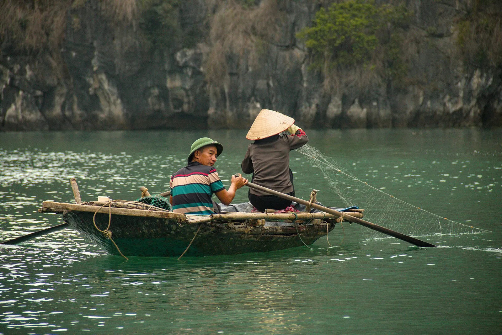Two people in conical hats in a small green rowing boat handling a fishing net in Bai Tu Long Bay