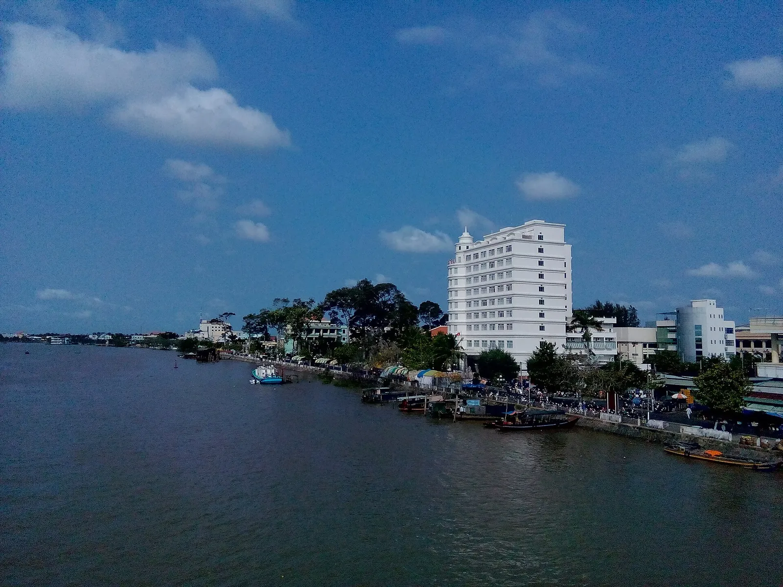 Ben Tre town seen from the river, with a white hotel tower and low buildings along the bank