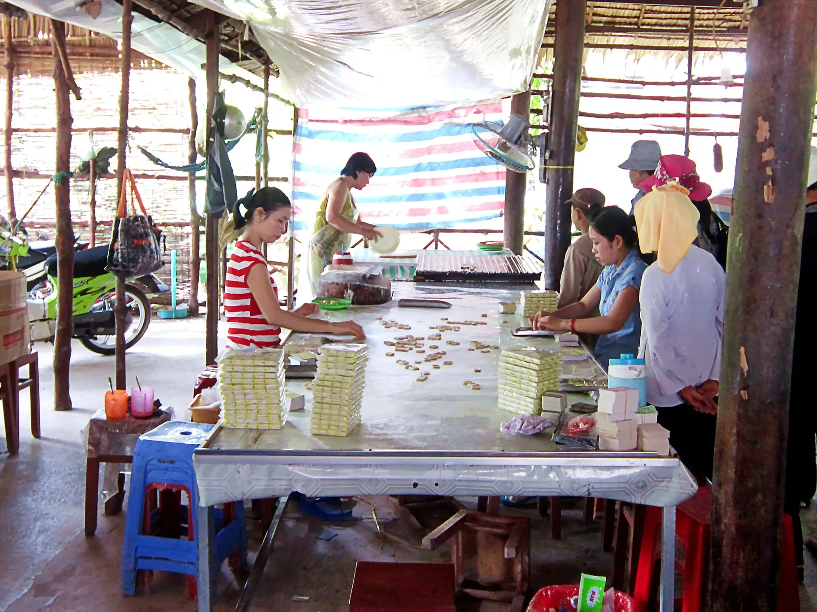 Workers cut and wrap coconut candy at long tables in an open-sided workshop