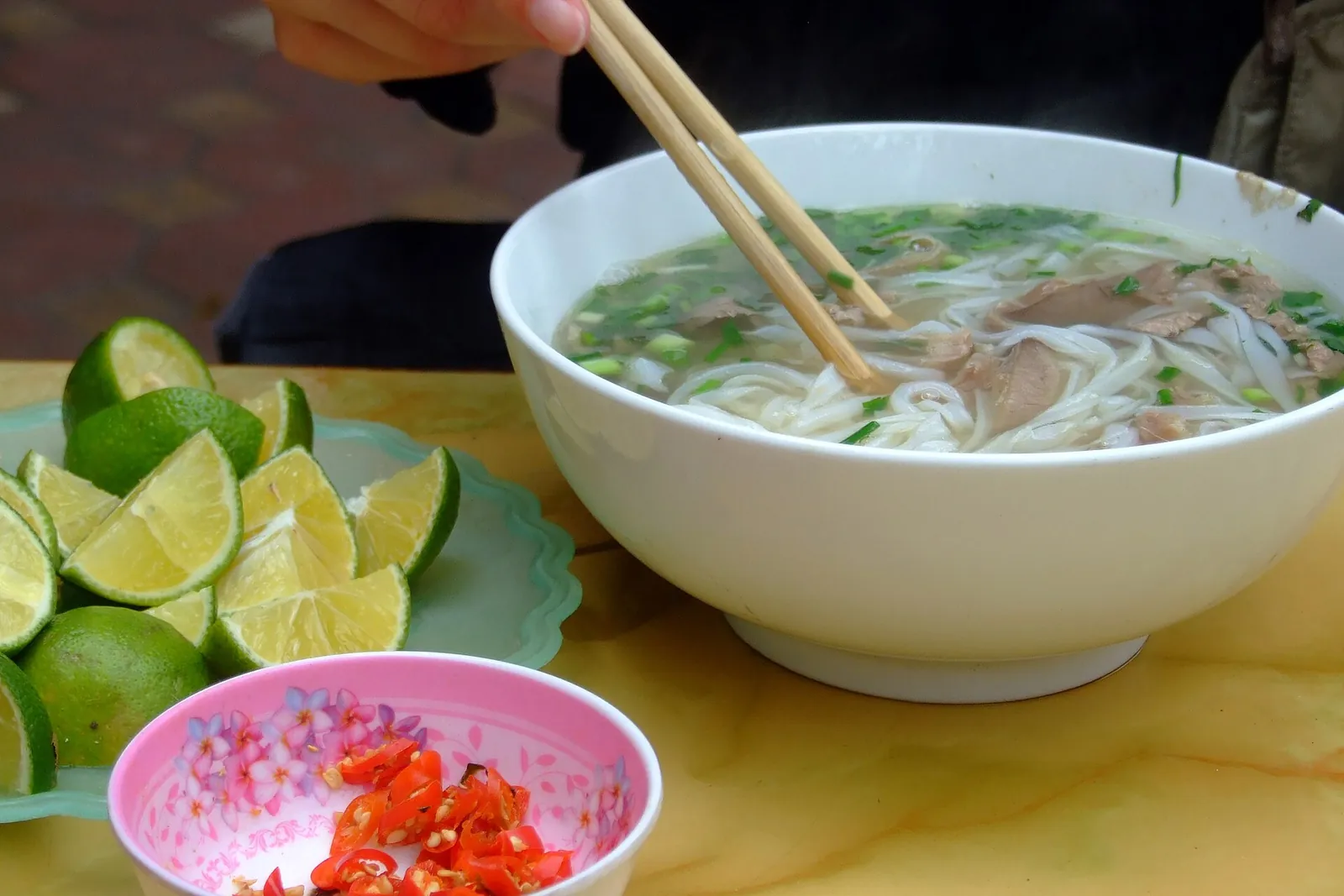 A bowl of beef pho with rice noodles, with lime wedges and sliced chilli on the side