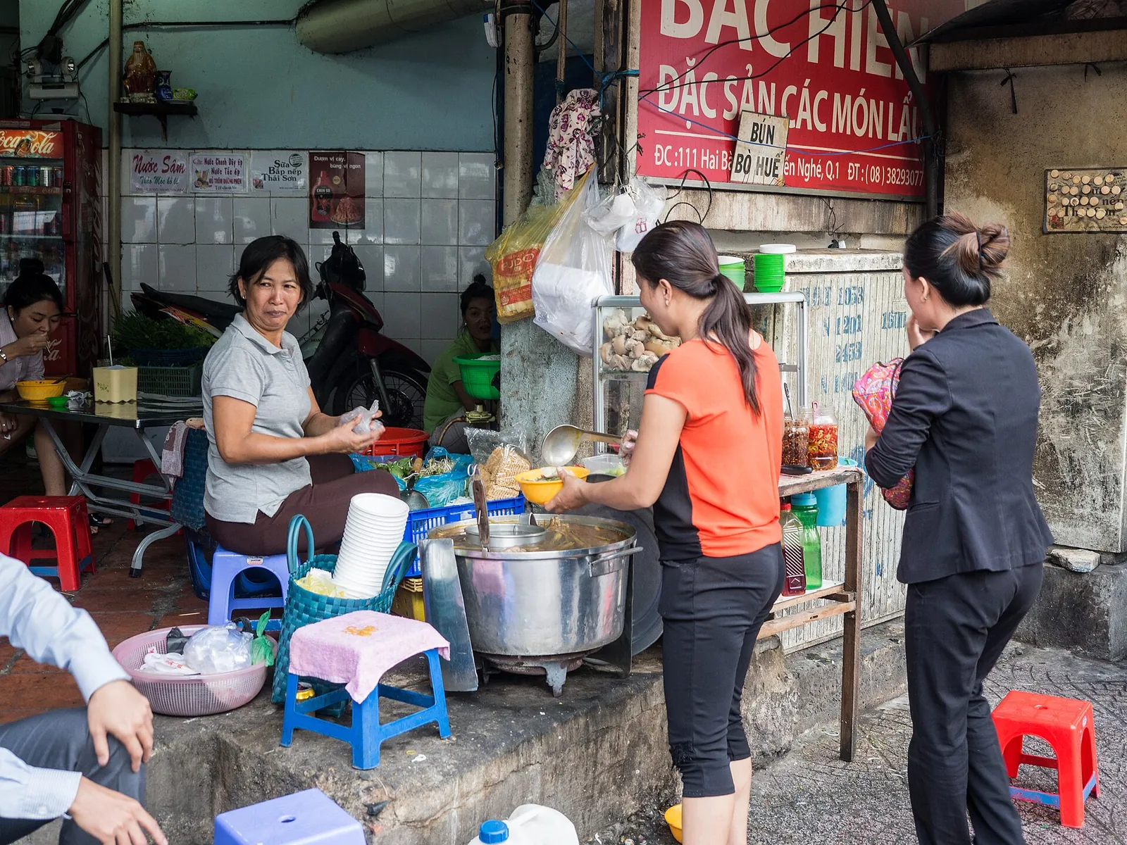 Pavement food stall in Hanoi with low plastic stools, a large soup pot and customers ordering