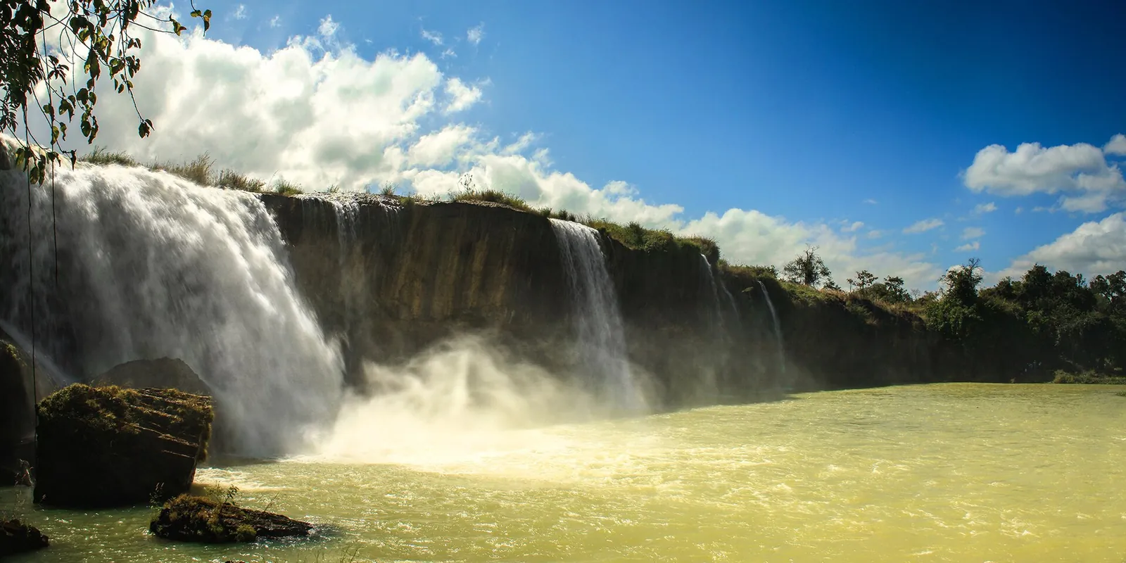 Dray Nur waterfall pouring over a wide basalt ledge into the Serepok River under a blue sky