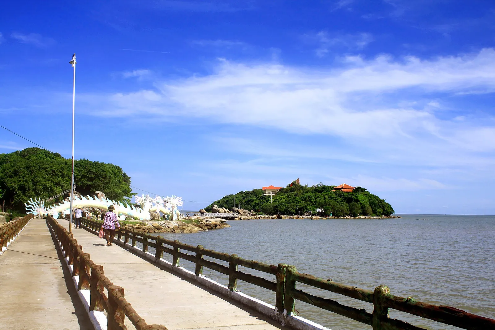 Concrete walkway with railings leading along the water towards the green islets of Hon Da Bac under a blue sky