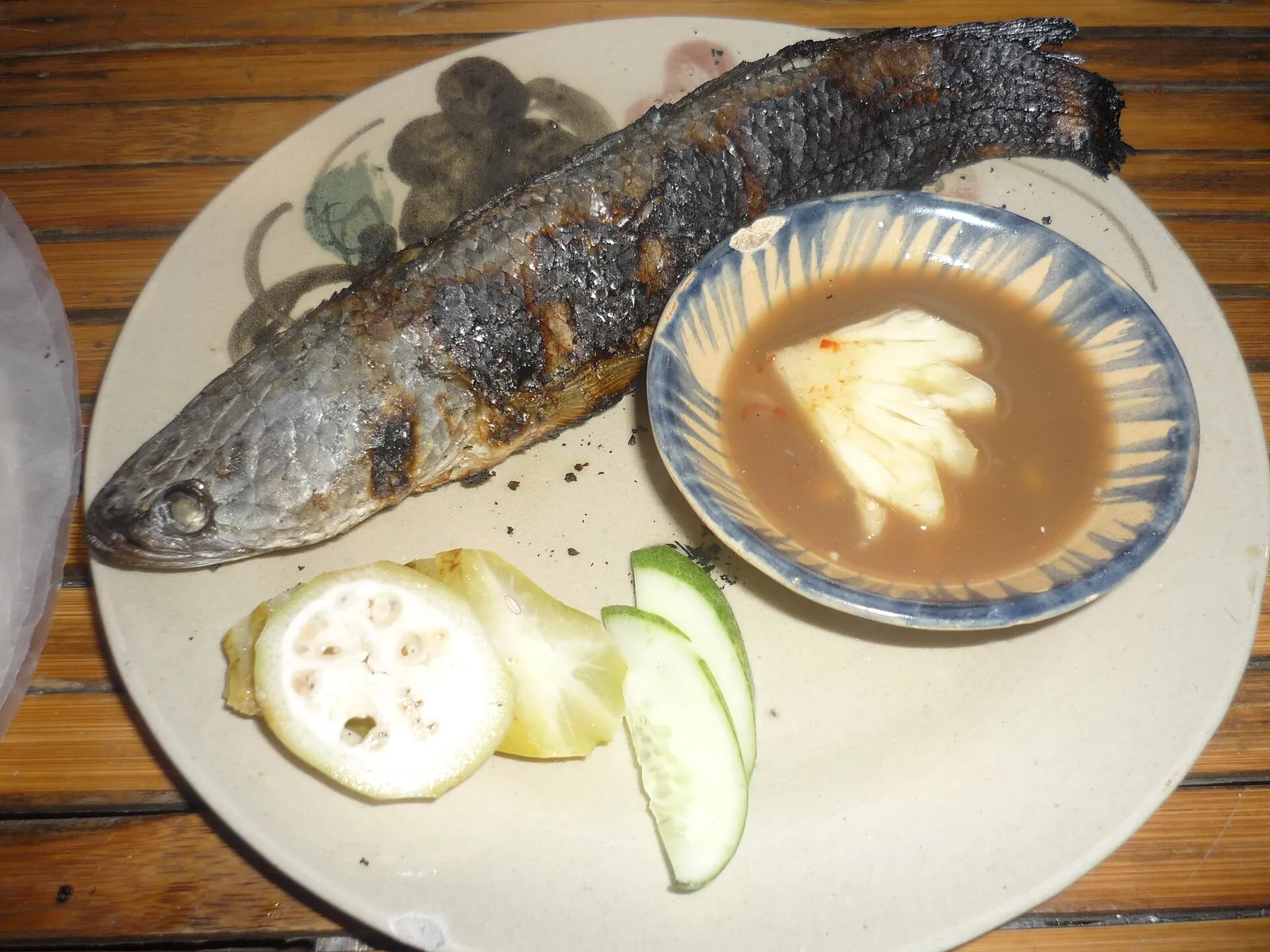 Whole grilled snakehead fish on a plate with a bowl of dipping sauce, cucumber and sliced green fruit