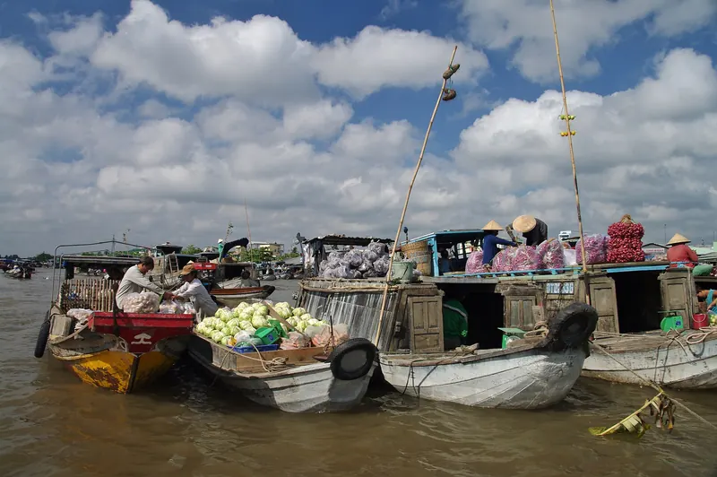 Wooden market boats loaded with fruit and vegetables moored together on the brown river under a blue, cloud-streaked sky at Cai Rang