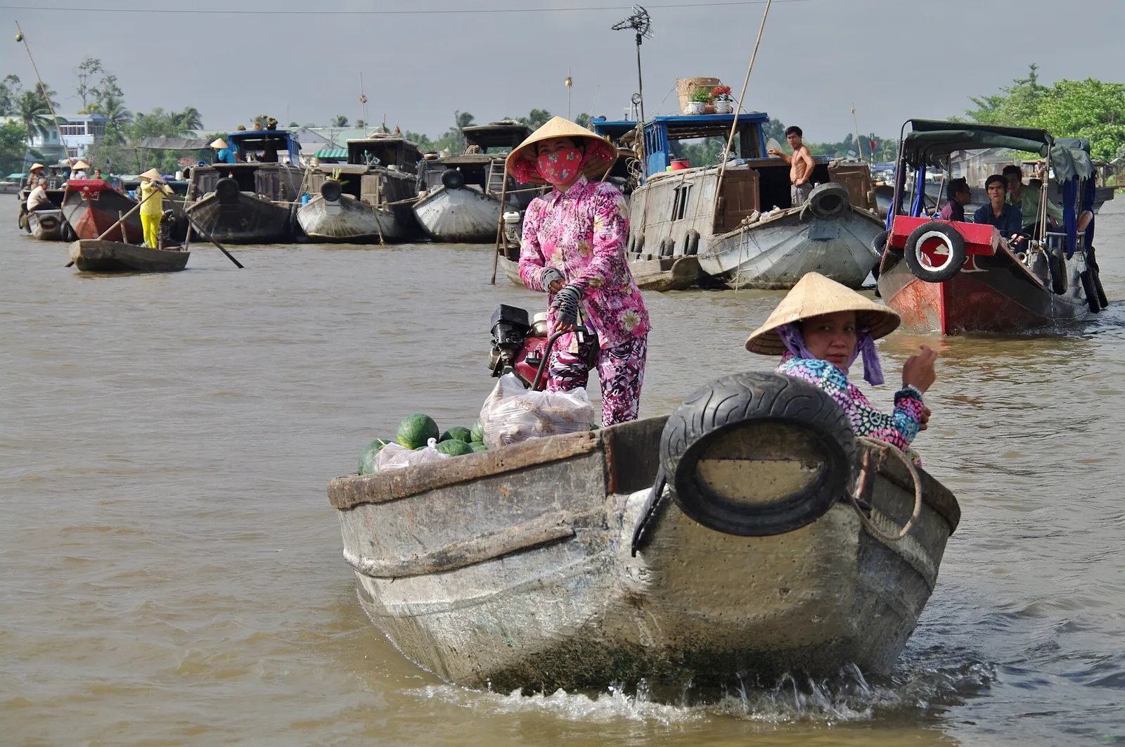 Two vendors in conical hats, one in pink, in a small loaded boat moving between larger trading boats on the river at Cai Rang