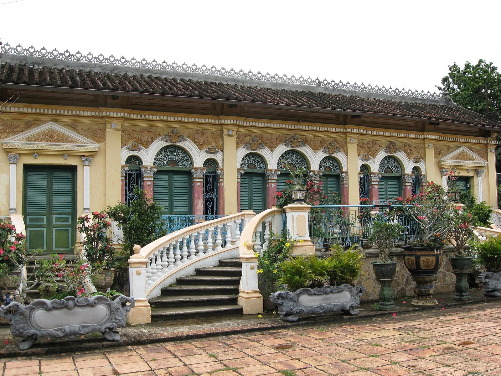 The yellow Binh Thuy ancient house with green shutters, arched windows and a curved white balustraded staircase, with potted plants in the courtyard