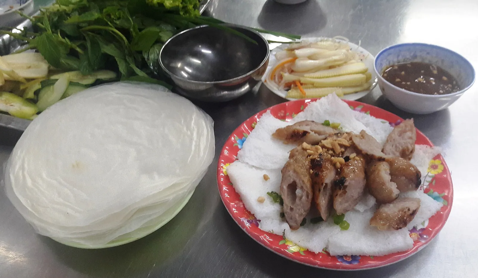 Grilled nem nướng pork on bánh hỏi rice vermicelli with a stack of rice paper, fresh greens, pickles and dipping sauce