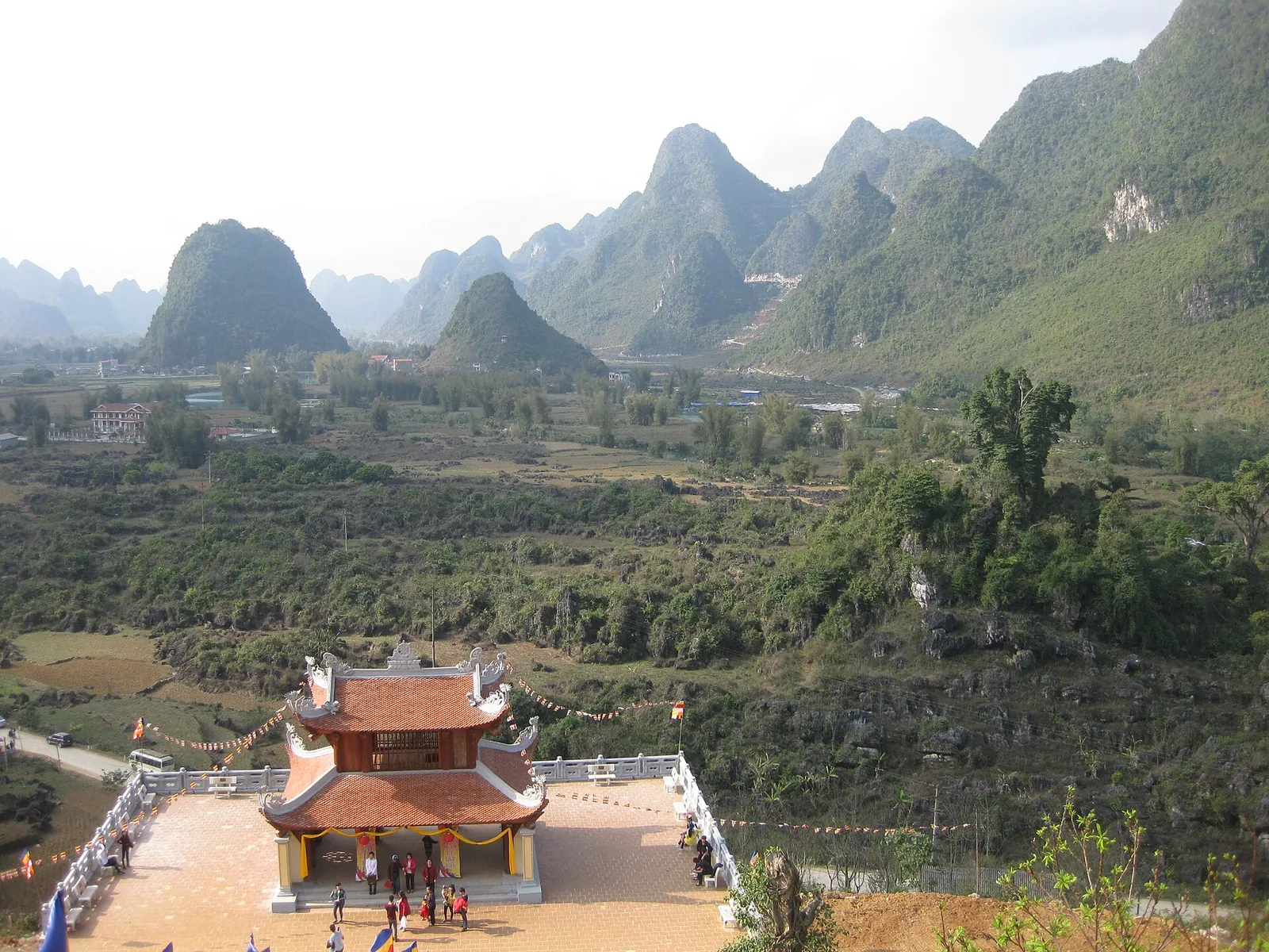 A red-roofed pagoda building on a hillside terrace overlooking a valley of limestone karst peaks near Ban Gioc