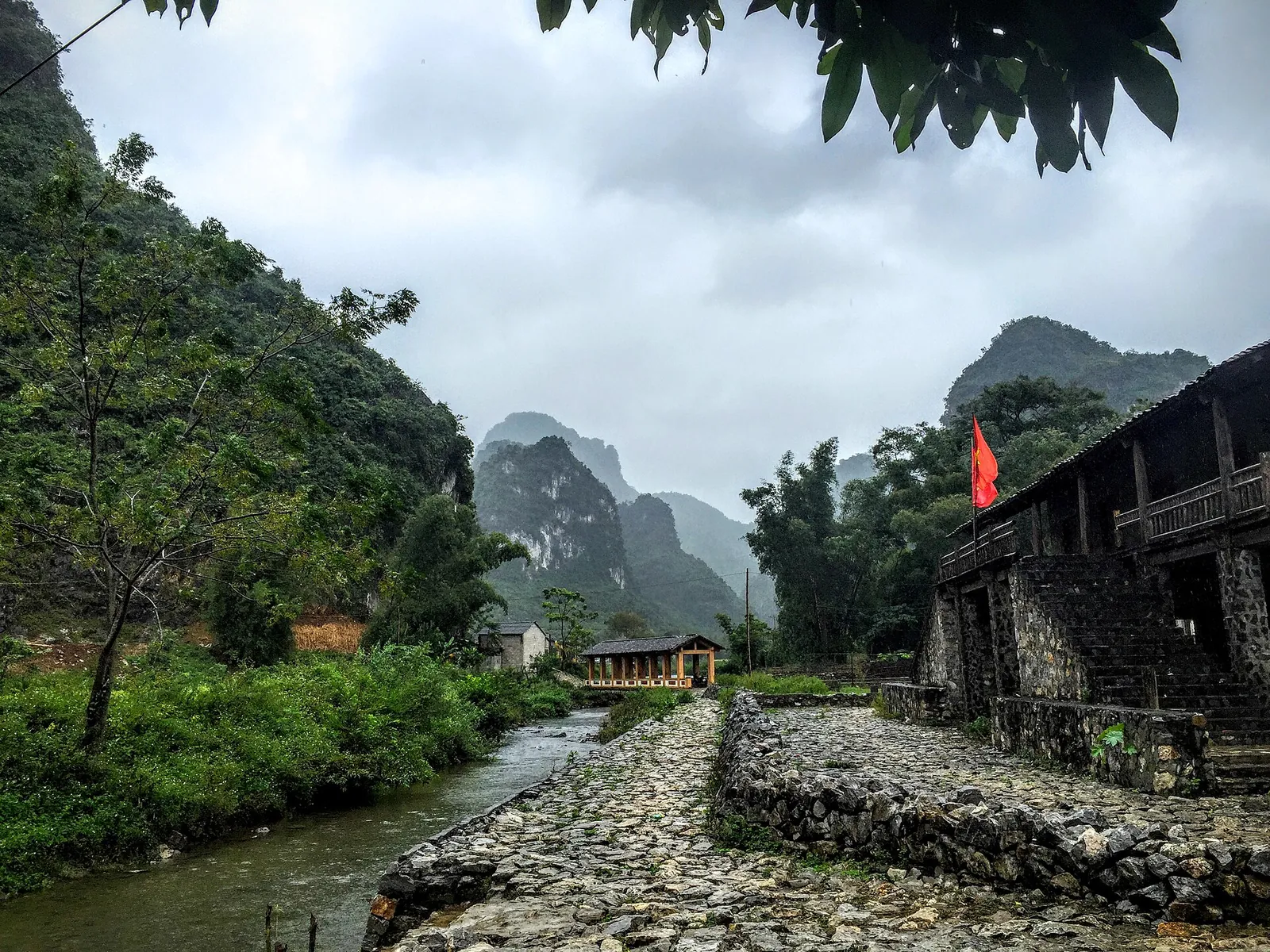 A stone-paved path beside a stream leading past stone houses in a Tay village under karst hills in Dam Thuy