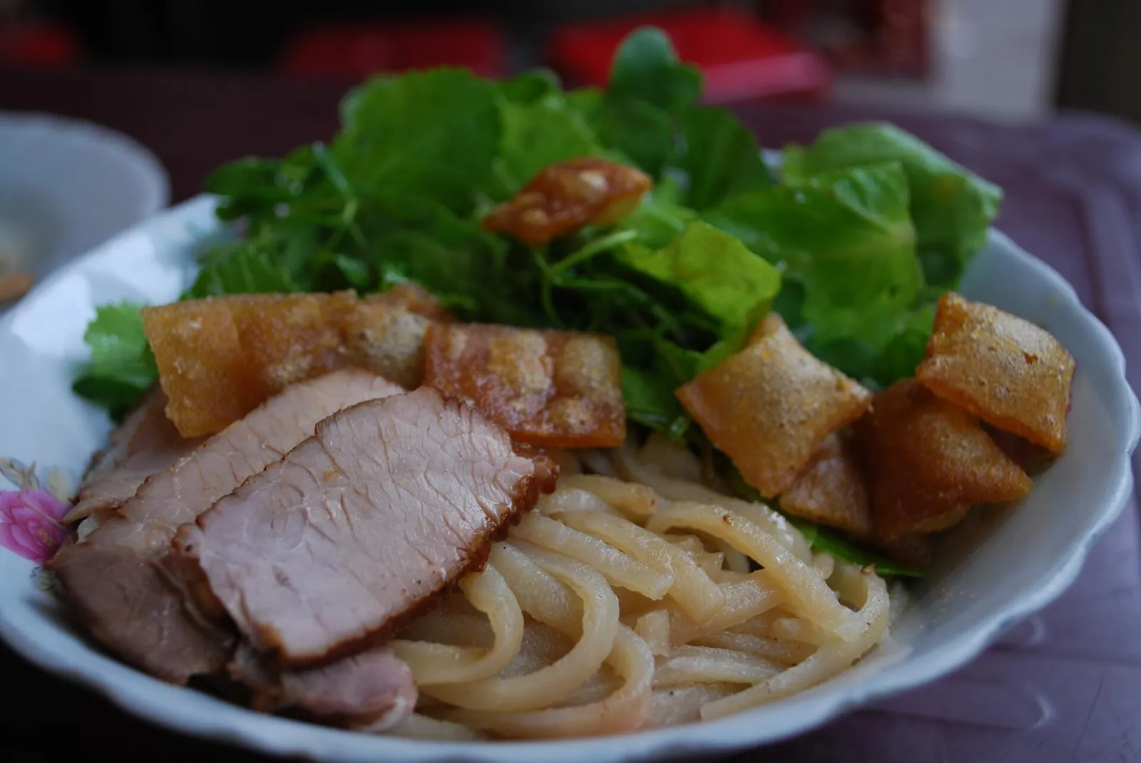A bowl of cao lau noodles topped with sliced pork, crisp crackers and fresh herbs