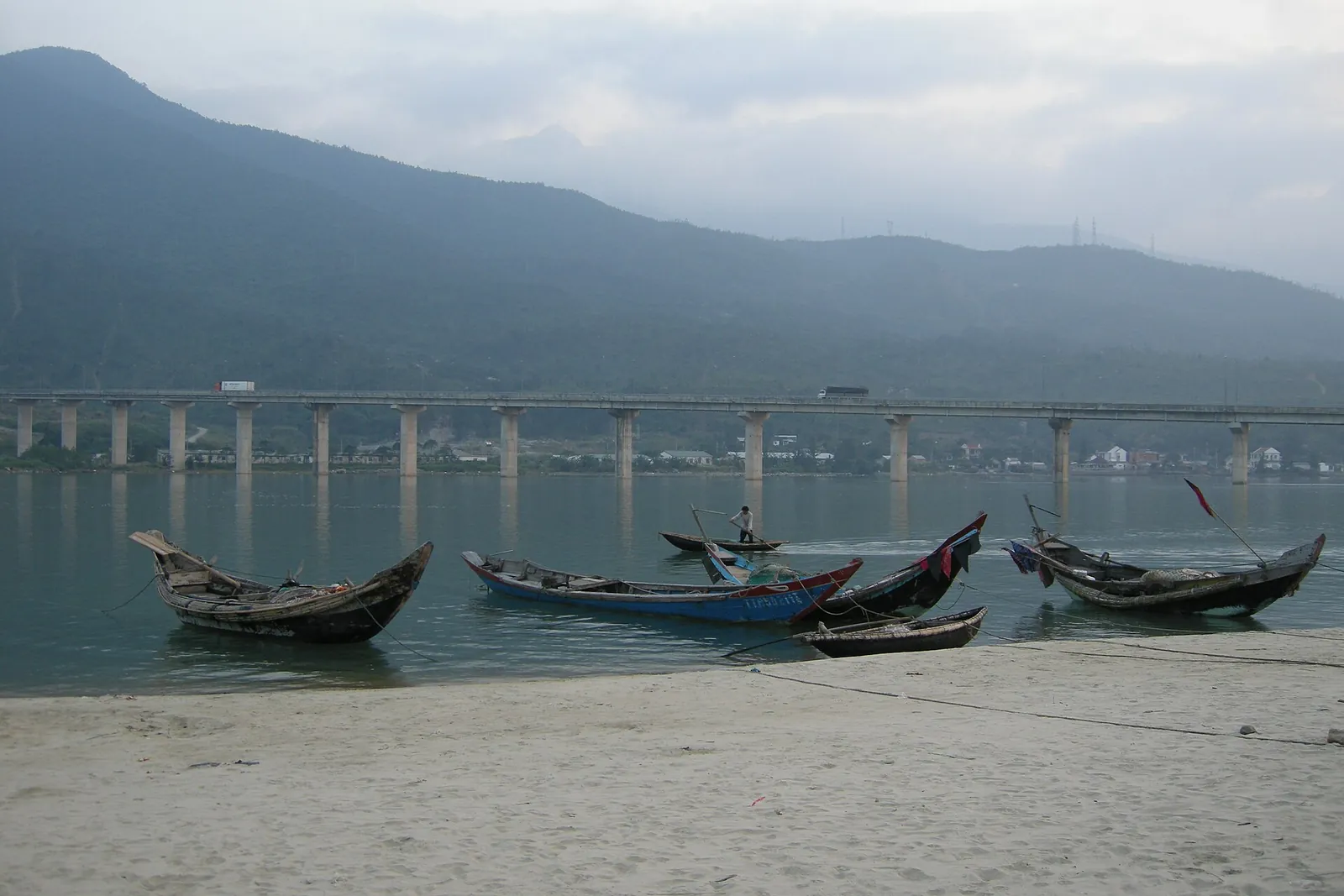 Wooden fishing boats pulled up on the shore of Lang Co lagoon, with a road bridge and mountains behind