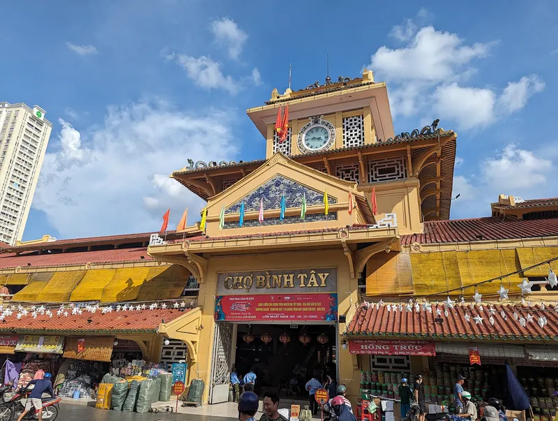 The yellow clock tower and front gate of Binh Tay Market under a blue sky, with shop awnings below