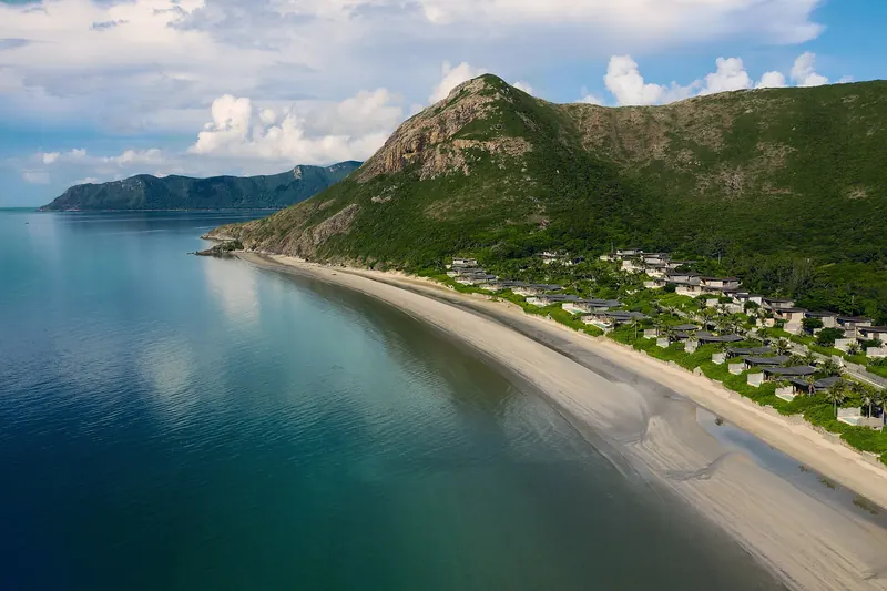 Aerial view of a curving sandy bay with villas along the shore below forested hills at Dat Doc on Con Son island