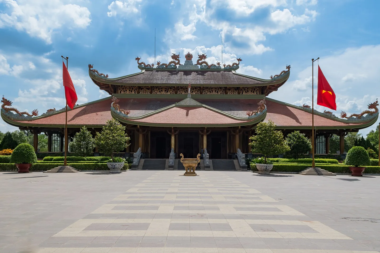 Main hall of the Ben Duoc Martyrs' Memorial Temple with curved tiled roofs, flags and a wide paved forecourt under a blue sky