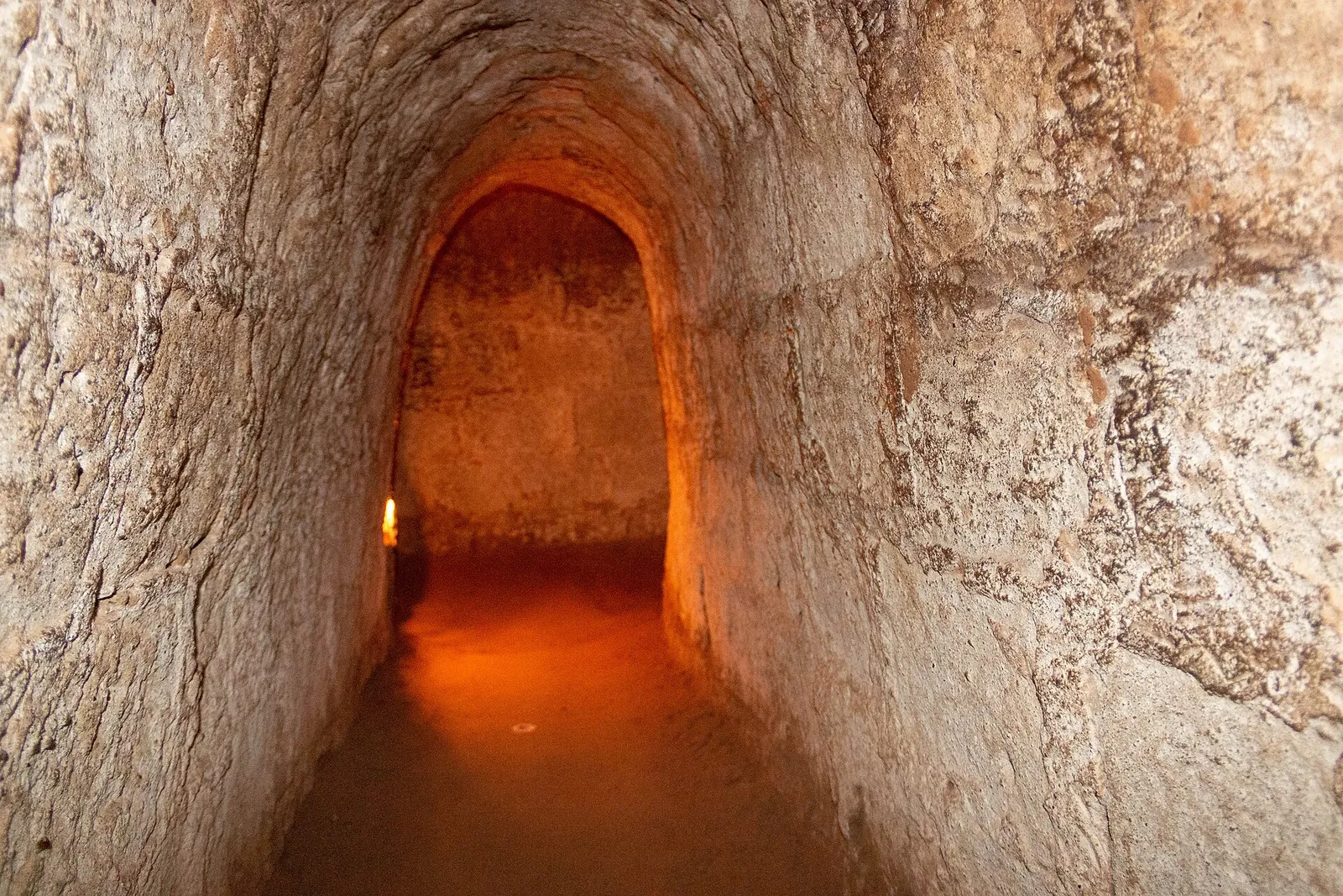 A narrow, lamp-lit tunnel passage with rounded walls of reddish-brown clay at the Cu Chi Tunnels