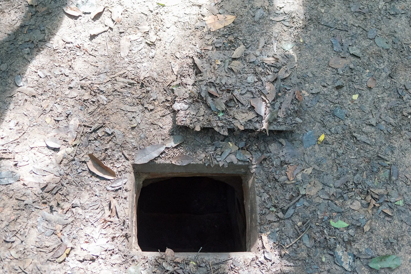 A small square tunnel entrance opening in leaf-covered ground at the Cu Chi Tunnels
