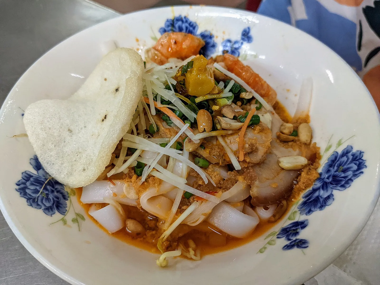 A bowl of mi Quang in Da Nang with flat rice noodles, pork, shrimp, peanuts, herbs and a rice cracker in a little broth