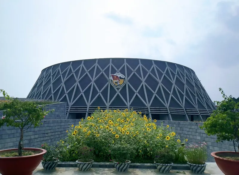 The hat-shaped Dien Bien Phu Victory Museum behind a bed of yellow flowers under a pale sky