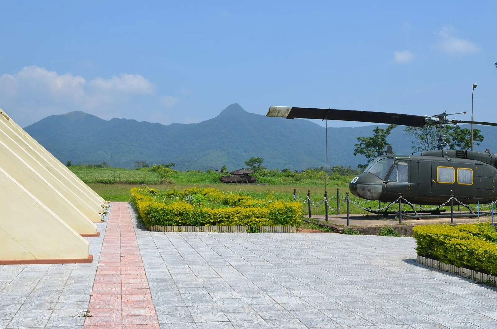 A Huey helicopter on display beside a paved path at the Khe Sanh base site, with green hills behind