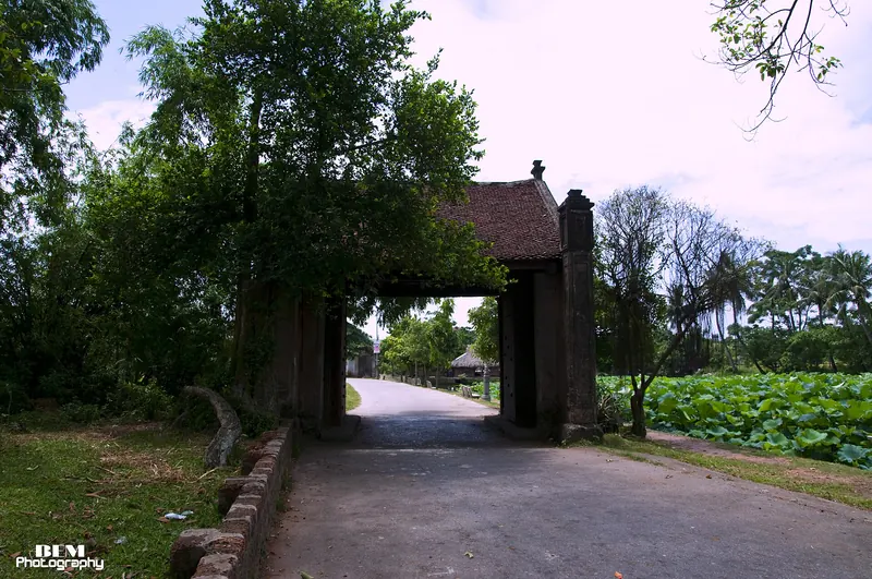 Tiled village gate over a lane in Duong Lam, with trees and a lotus pond beside it