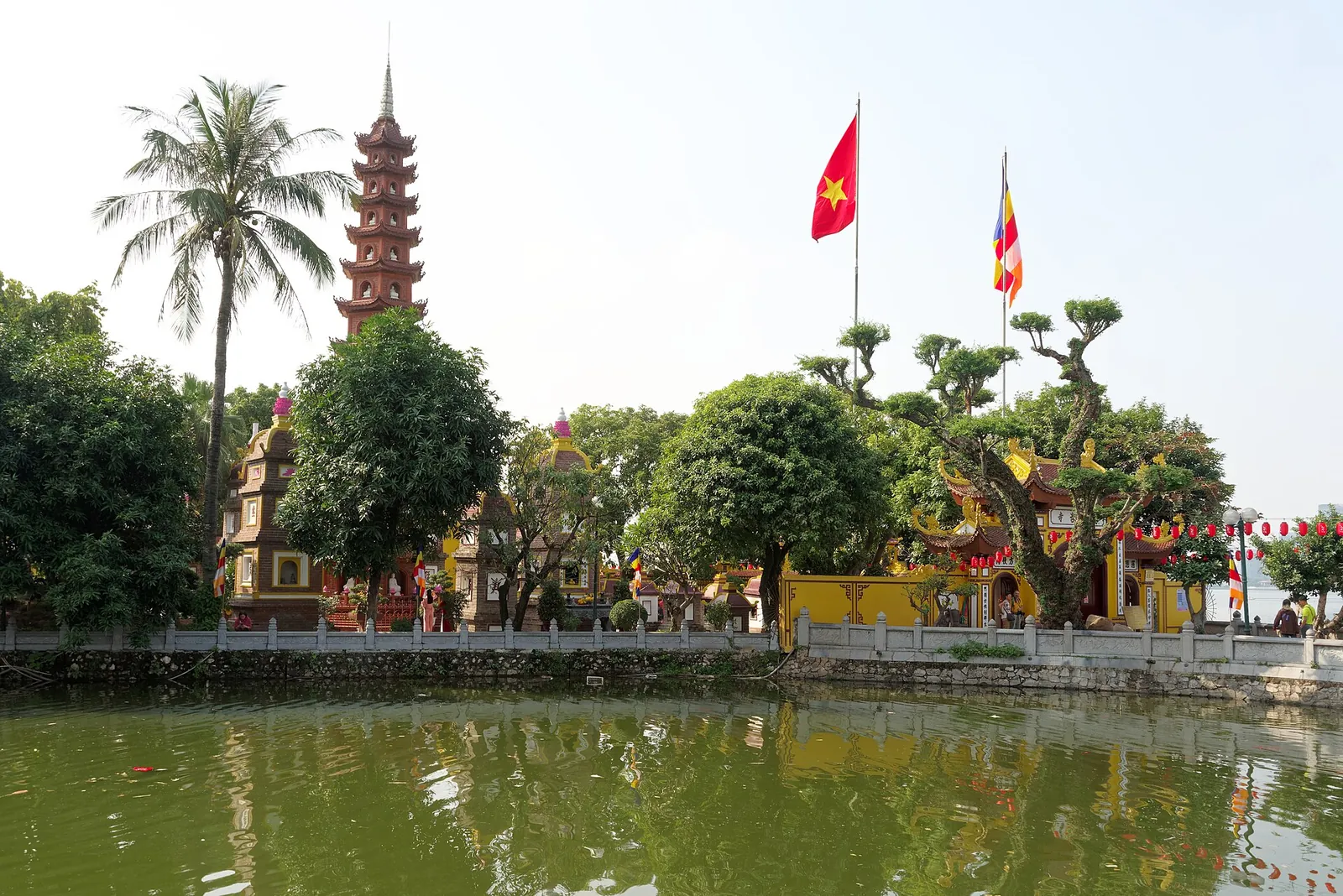 Tran Quoc Pagoda's red brick tower, yellow gate and flags among trees on the shore of West Lake, Hanoi