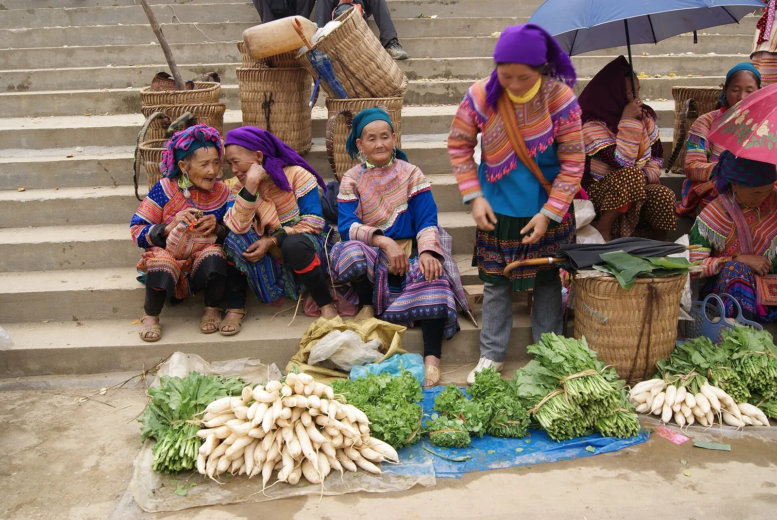 Hmong women in bright embroidered dress sitting on steps behind vegetables laid out for sale at Bac Ha Sunday market