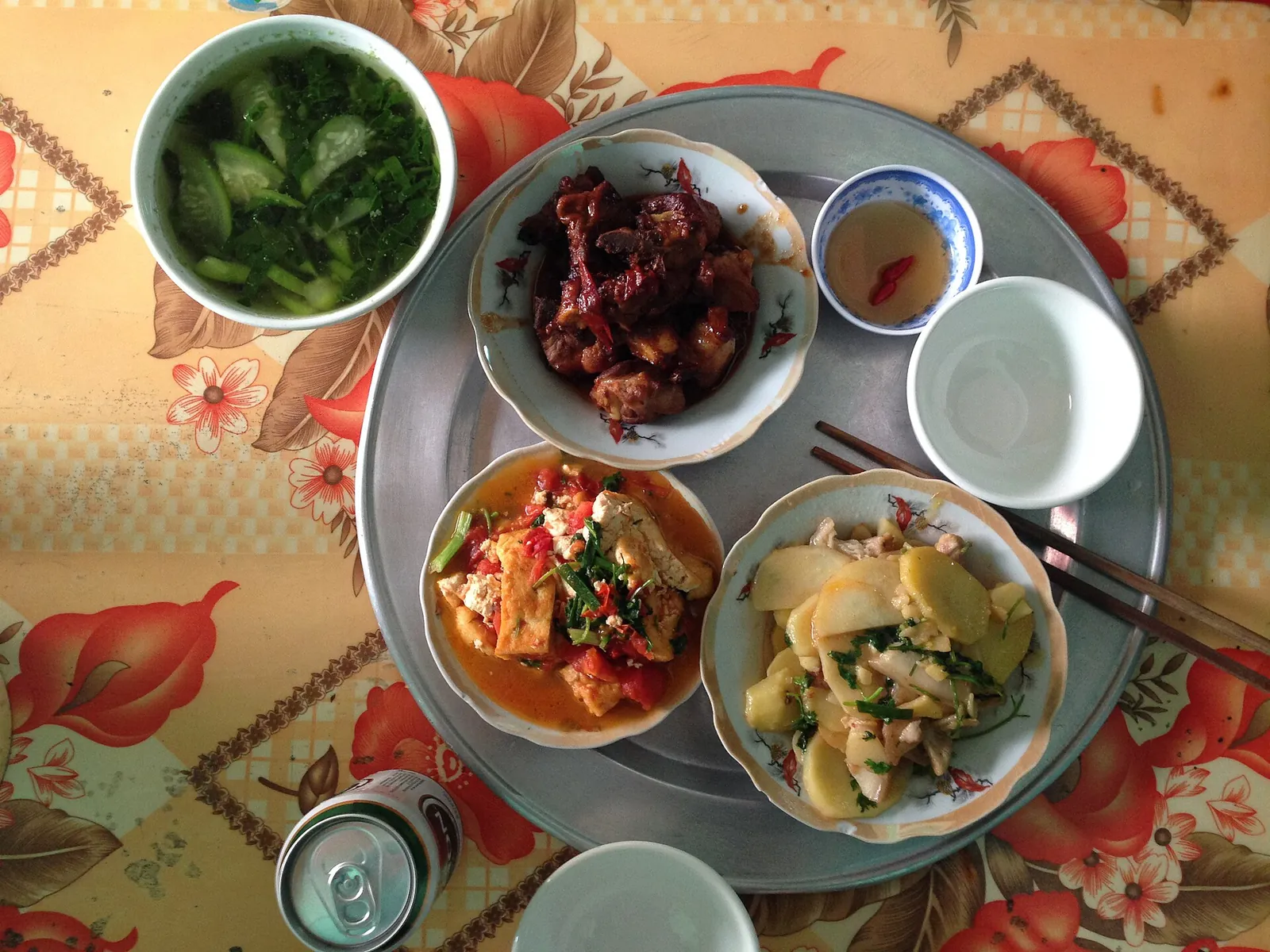 A Vietnamese home meal on a round aluminium tray: greens soup, sweet and sour pork ribs, stir-fried potatoes, tofu in tomato sauce and a small bowl of fish sauce, with chopsticks