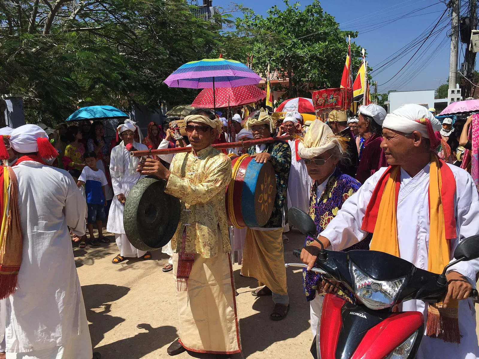 Cham men in white robes and turbans with gongs and drums walking in a Kate festival procession under umbrellas