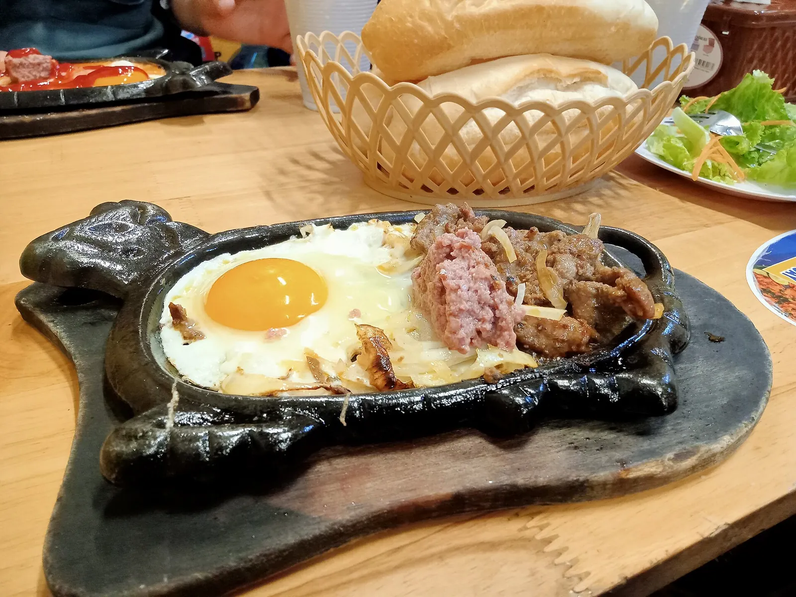 Bánh mì baguette in a basket beside a sizzling iron plate of beef, egg and pâté