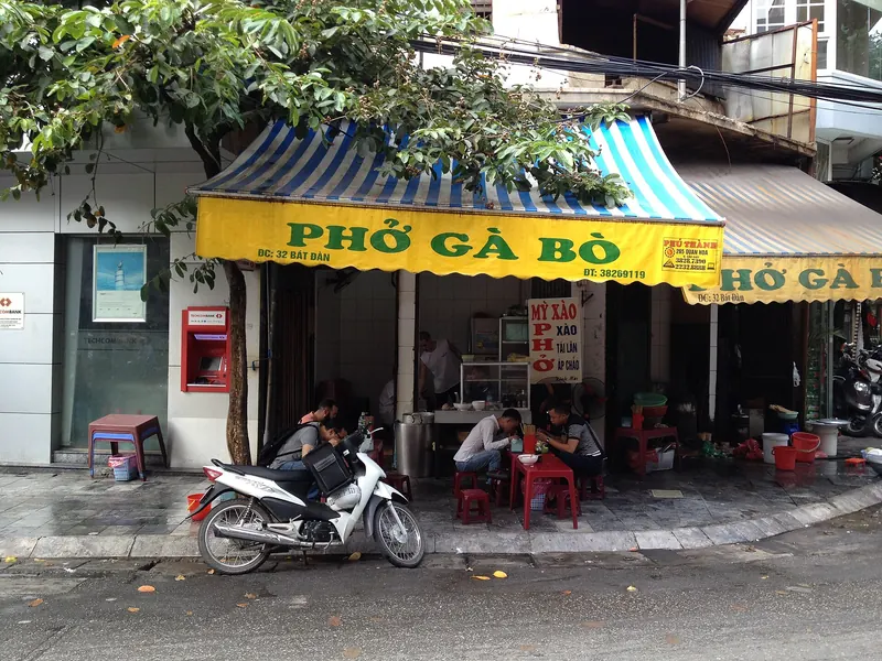Phở shop at 32 Bát Đàn street in Hanoi, with a striped awning, plastic stools and a parked motorbike out front