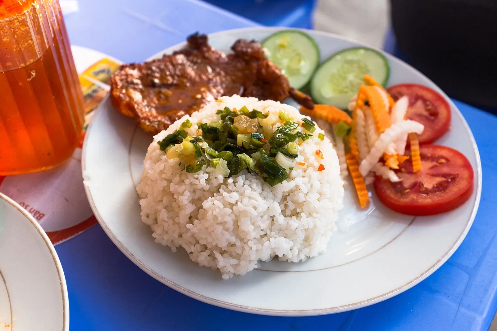 A plate of cơm tấm with grilled pork, rice topped with spring onion, tomato, cucumber and pickled carrot