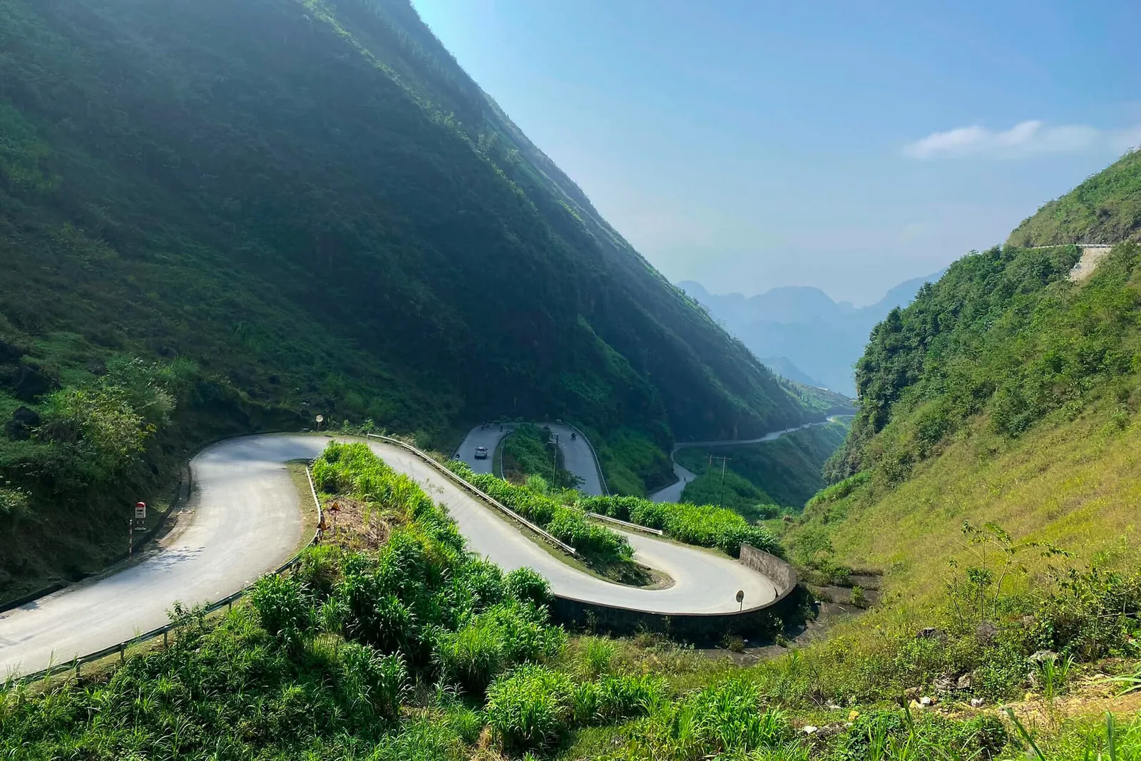 A road looping in tight hairpin bends down a green mountainside under a blue sky