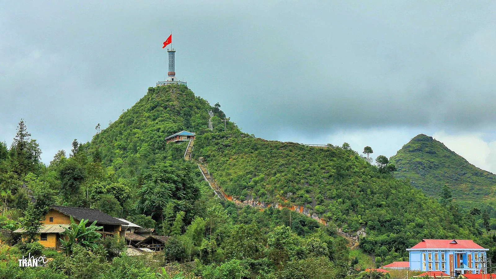 A large red flag flying from a tower on a green hilltop above village houses