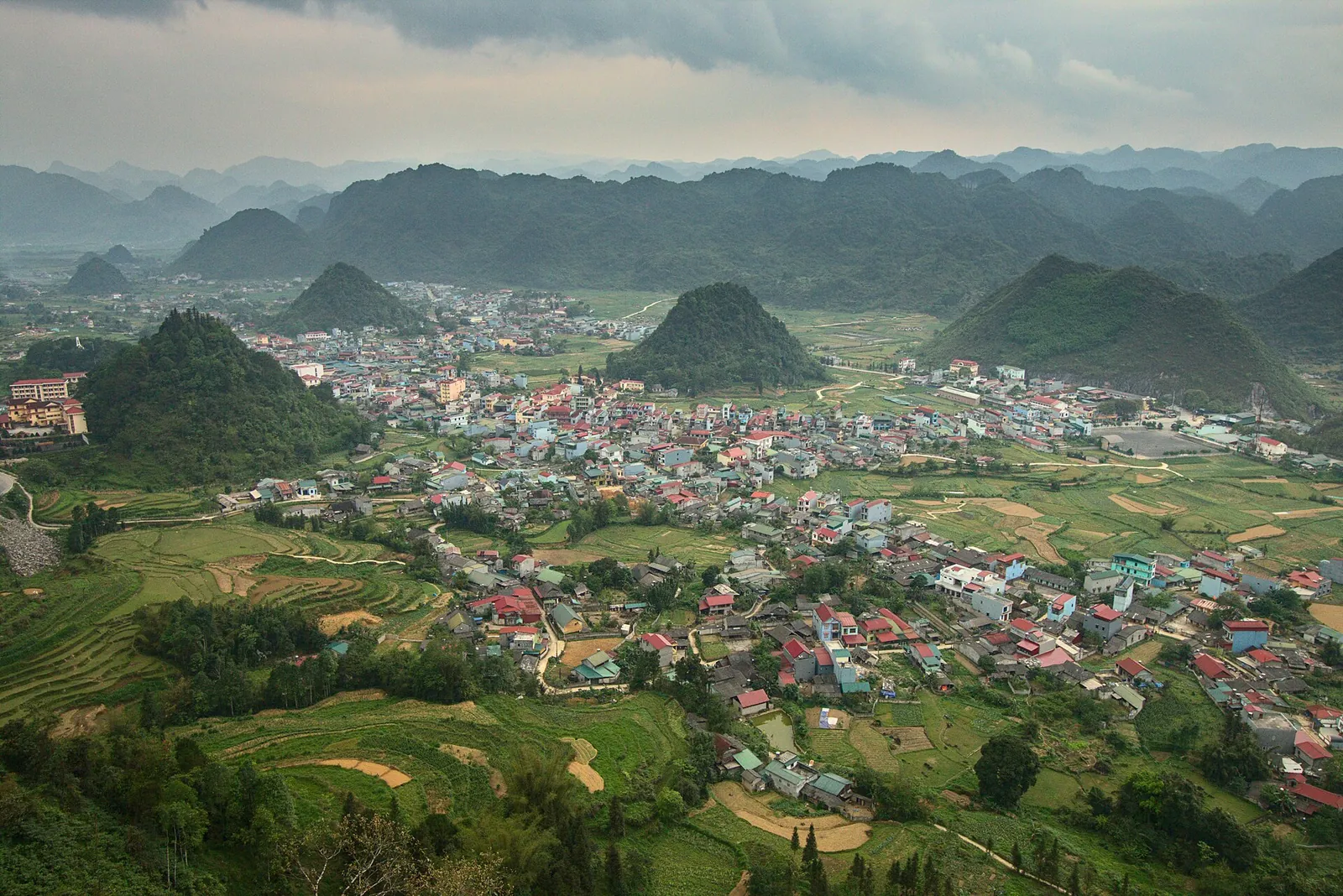 A wide green valley with a town spread across its floor and rows of conical limestone hills behind, in Quan Ba