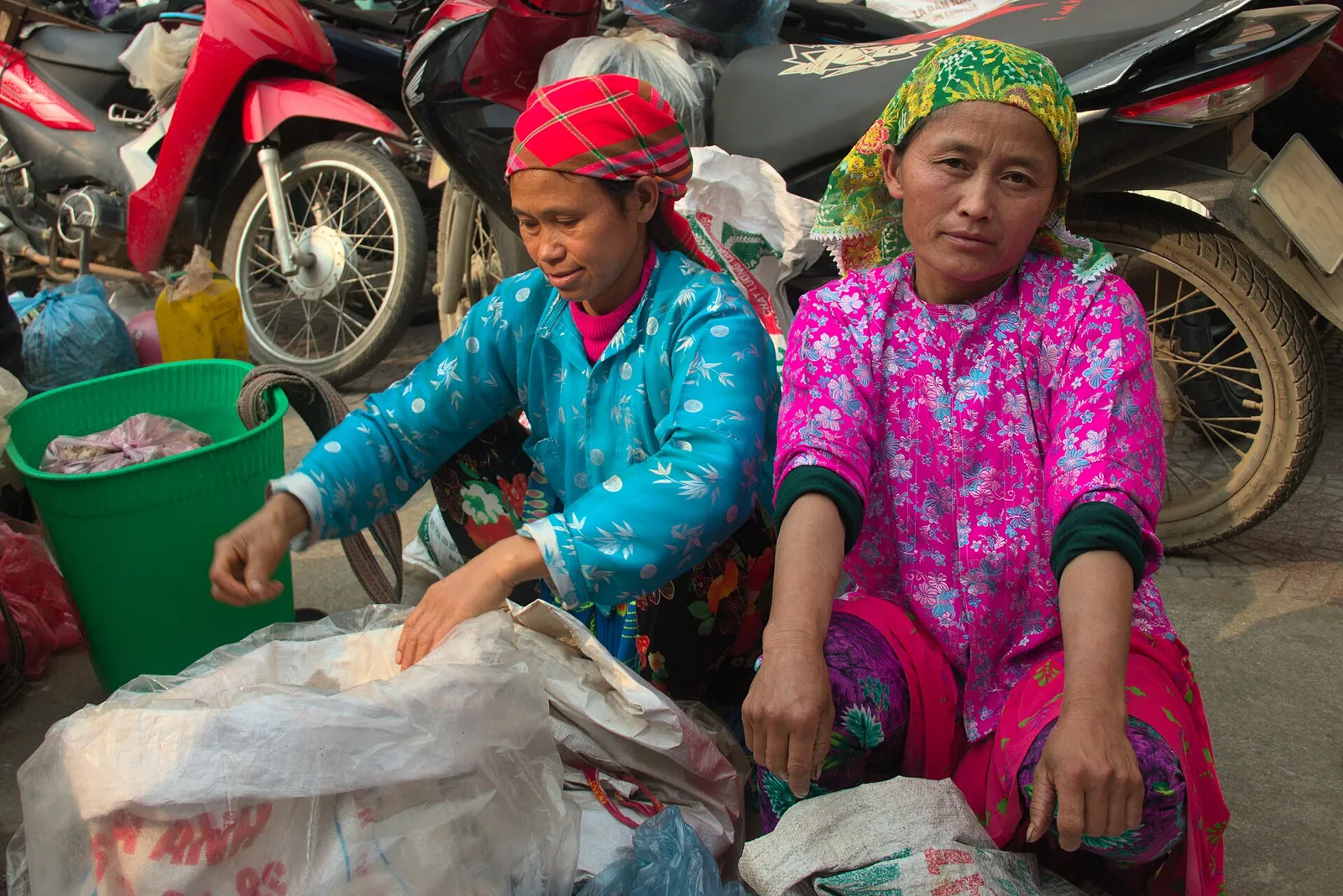 Two women in bright pink and blue traditional clothing sitting beside parked motorbikes at Meo Vac market