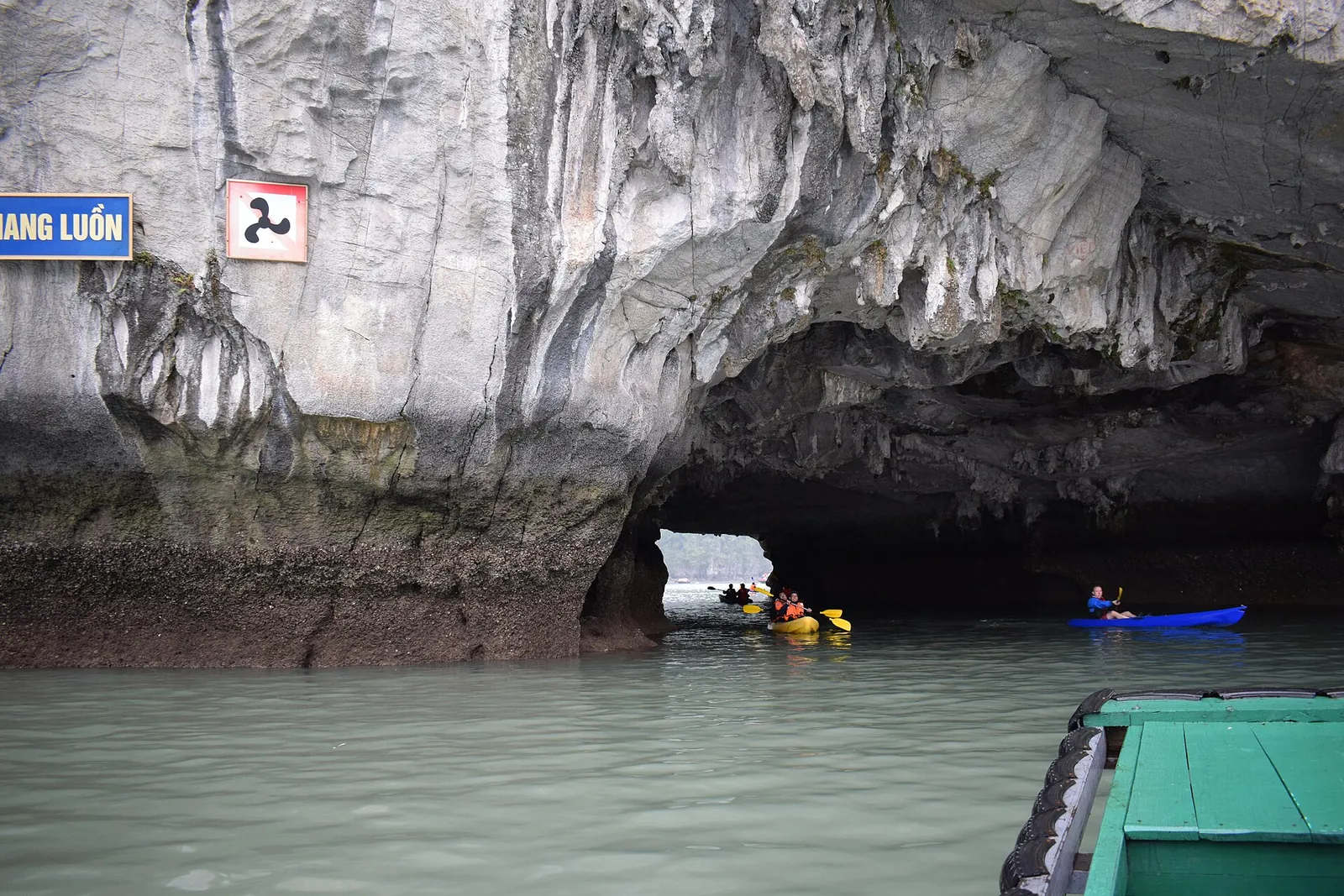 Kayakers paddling through the low rock arch of Luon Cave in Ha Long Bay, beside a moored tourist boat