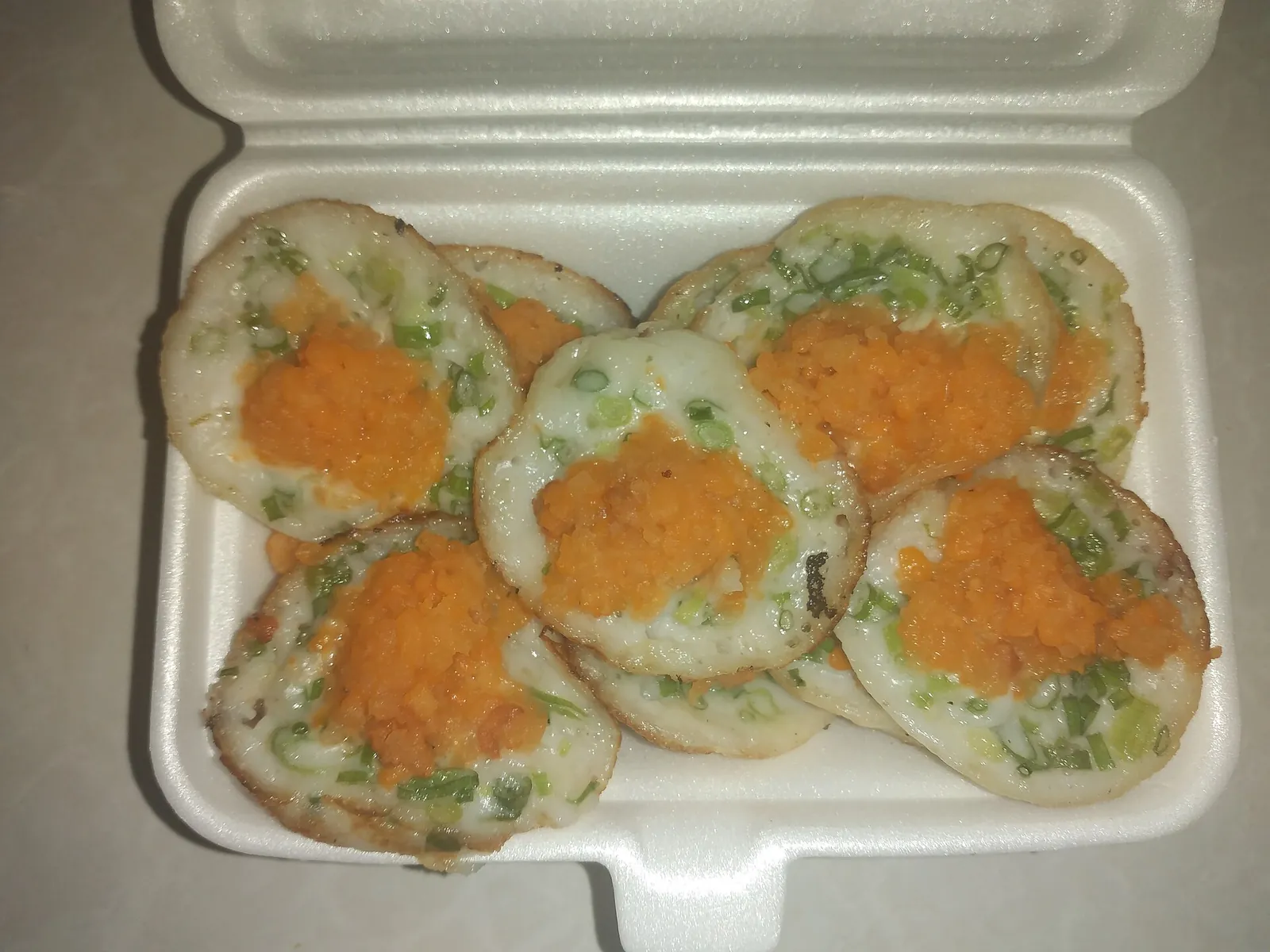 A takeaway box of bánh khọt, small round rice-flour pancakes topped with spring onion and orange shrimp floss