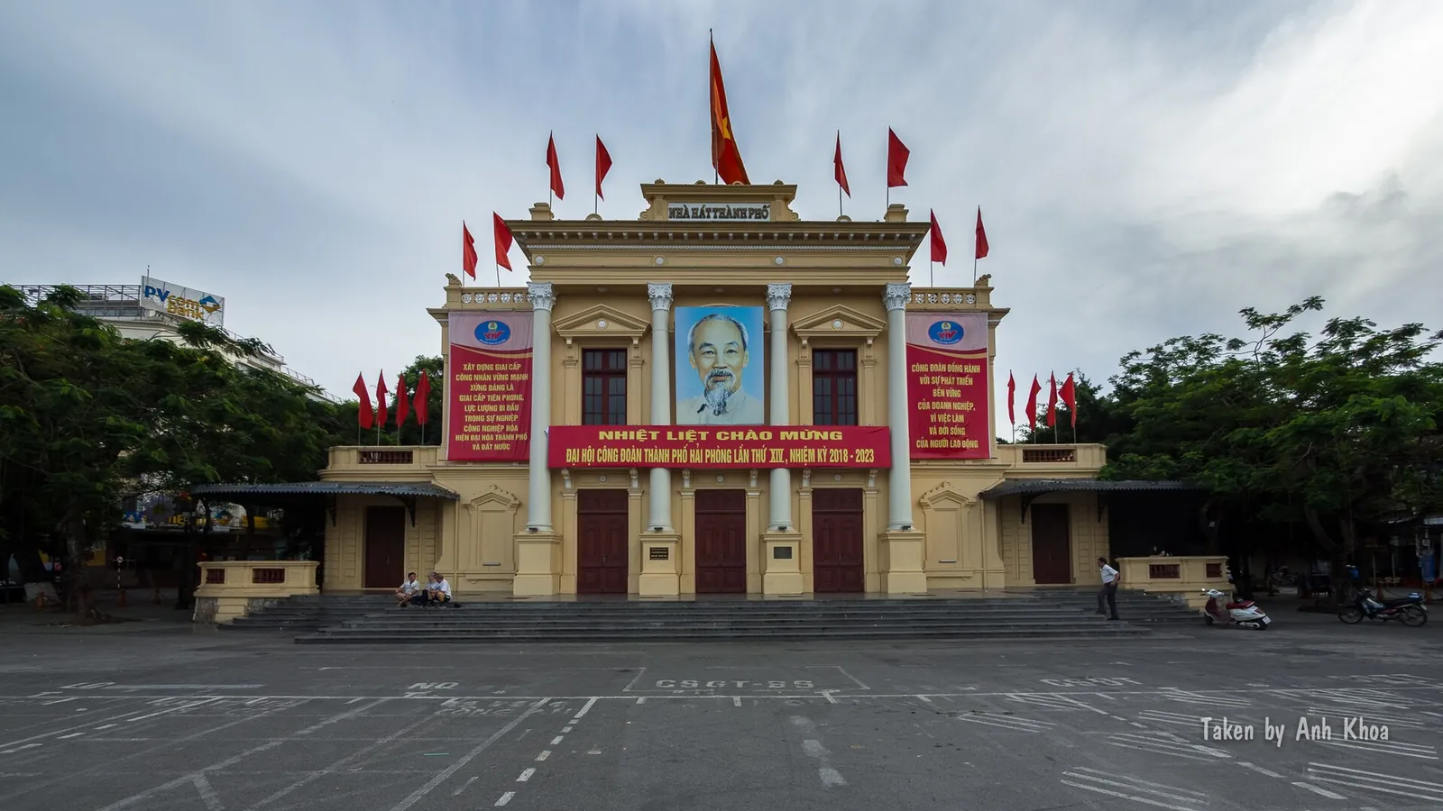 The yellow Hai Phong Opera House with red flags and banners along its roofline, seen across the open square in front of it