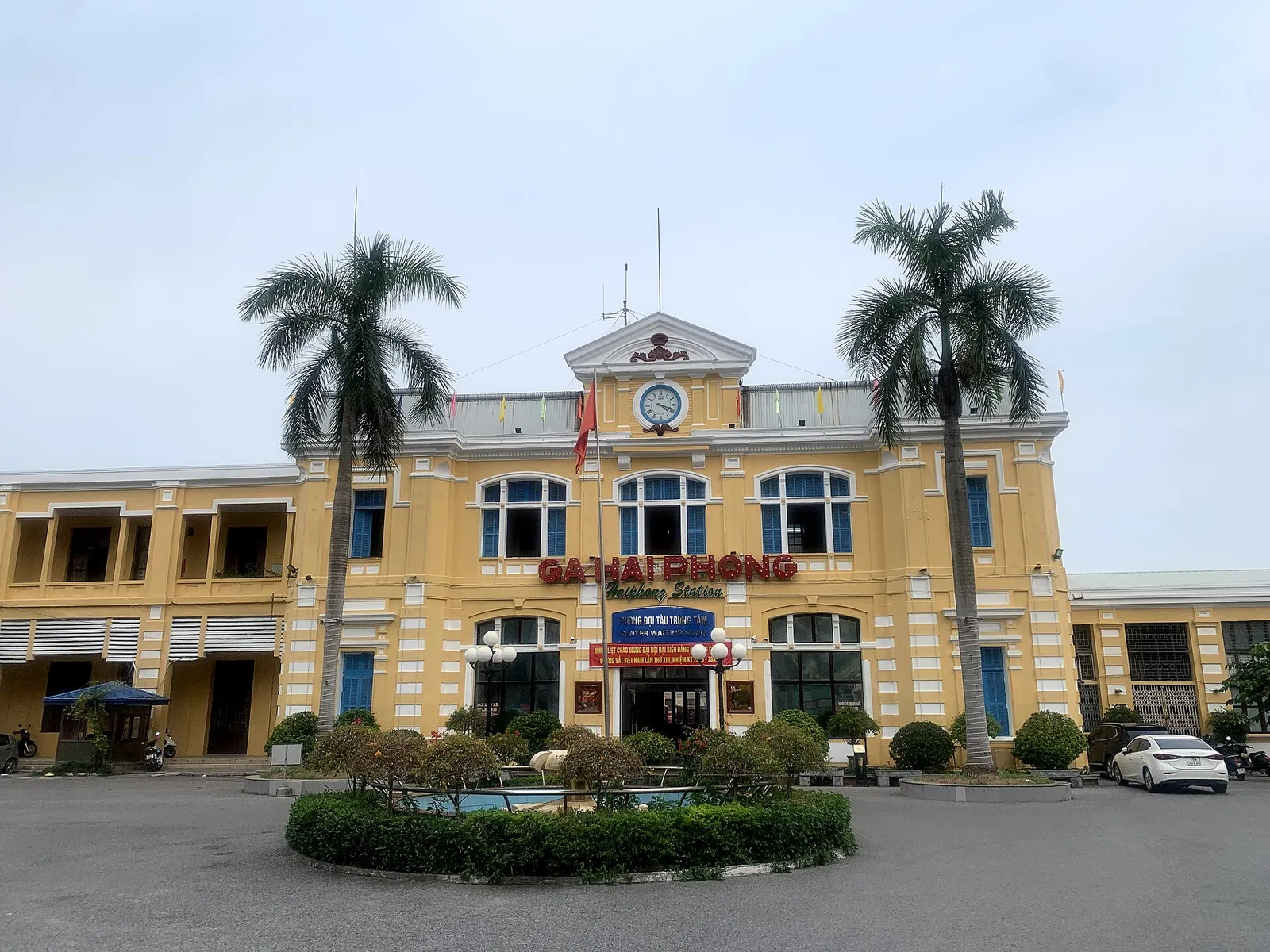 The yellow two-storey Hai Phong railway station with a clock above the entrance, palm trees and a round flowerbed in front
