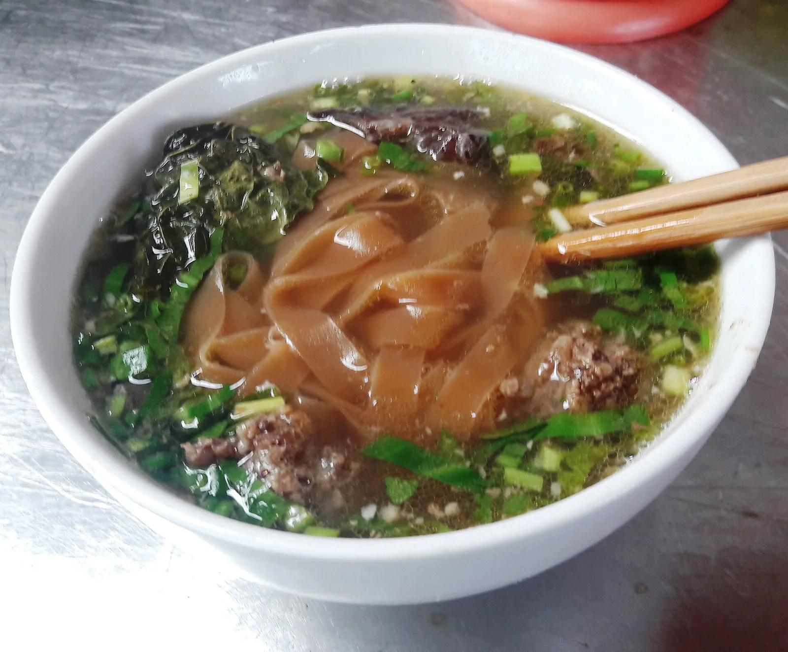 A bowl of banh da cua with flat reddish-brown rice noodles, crab broth, greens and spring onions, chopsticks resting on the rim