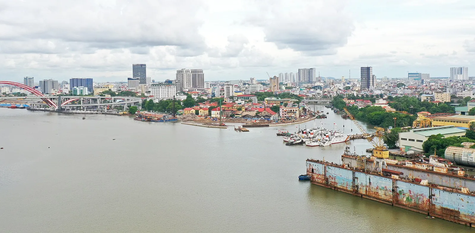 Wide view over the Tam Bac river where it meets a larger river, with boats moored along the bank and the Hai Phong skyline behind