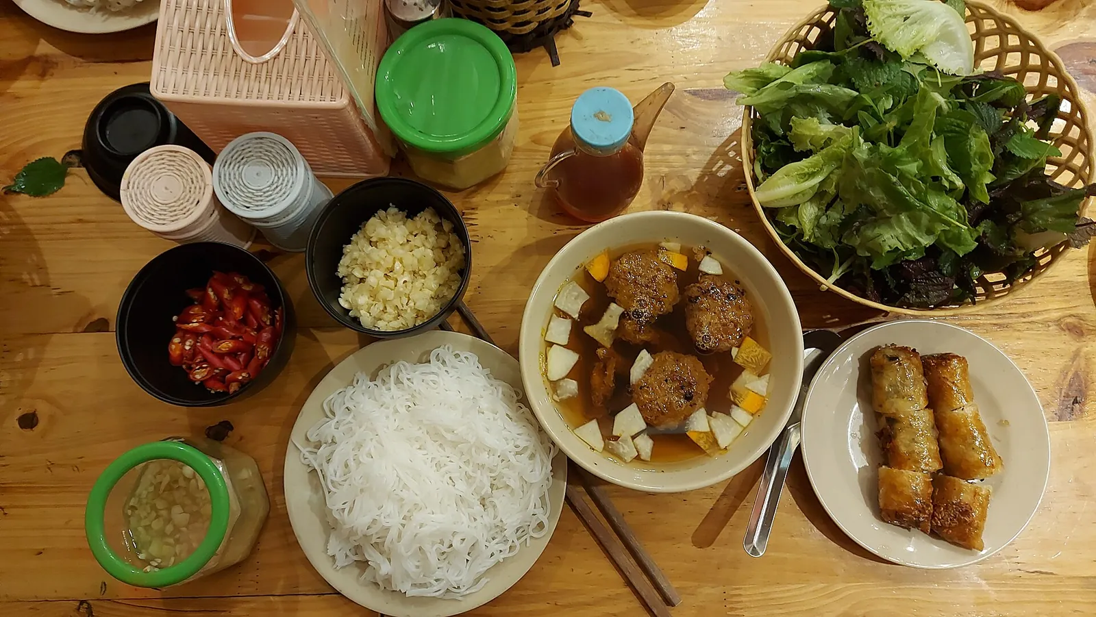 Bun cha laid out on a table: grilled pork in broth, rice vermicelli, fresh herbs, spring rolls, chilli and garlic