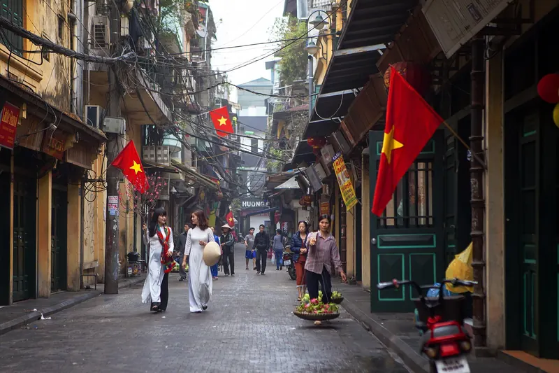 Ta Hien Street in Hanoi's Old Quarter by day, with yellow shophouses, Vietnamese flags and women in ao dai walking down the lane