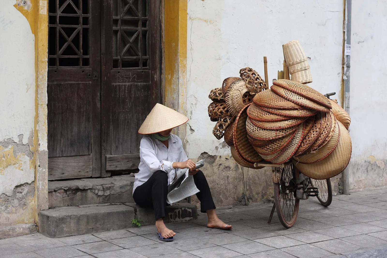 A street vendor in a conical hat resting beside a bicycle loaded with baskets in Hanoi's Old Quarter