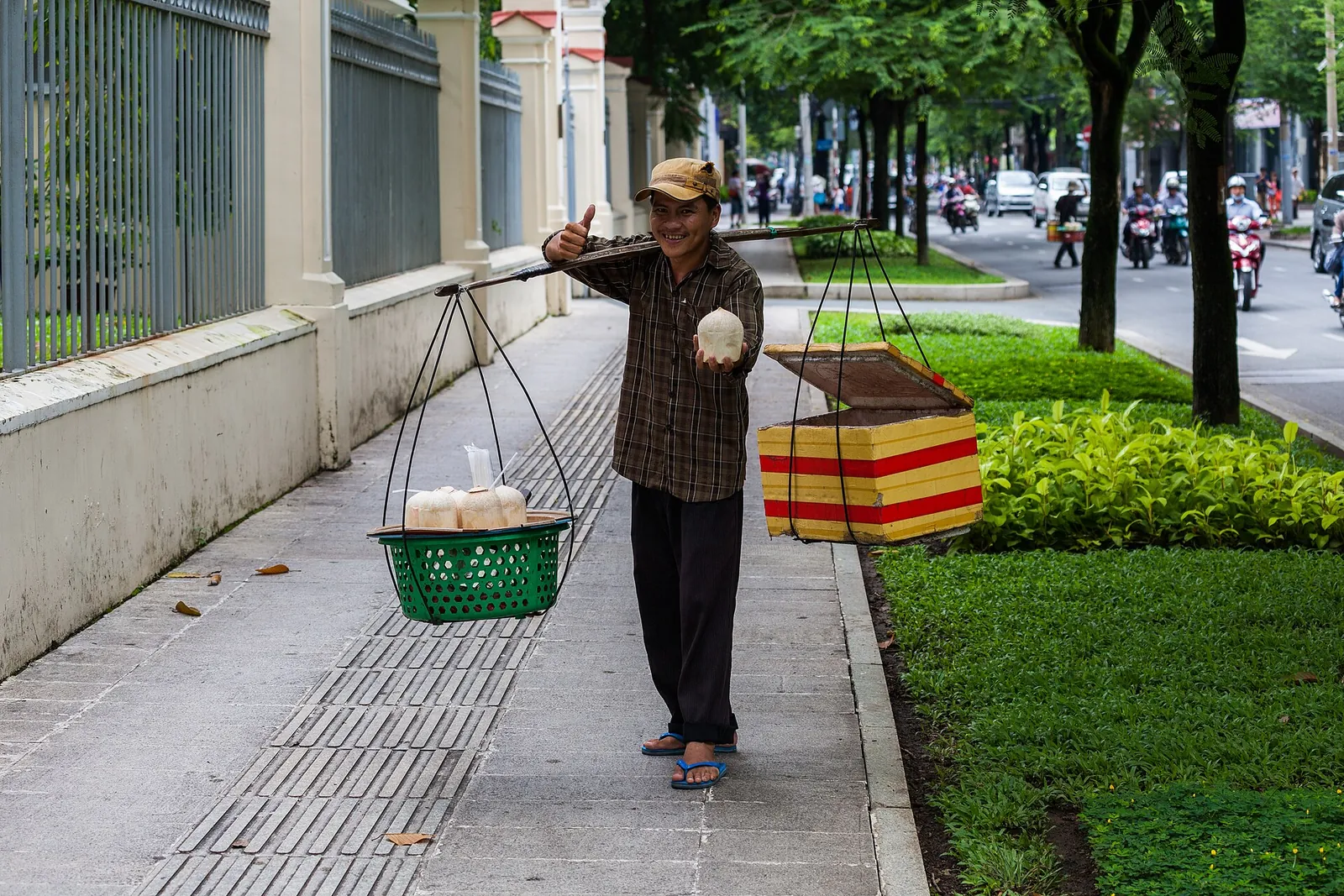 A street vendor in Ho Chi Minh City holding up a green coconut, with baskets of coconuts on a shoulder pole on a tree-lined pavement